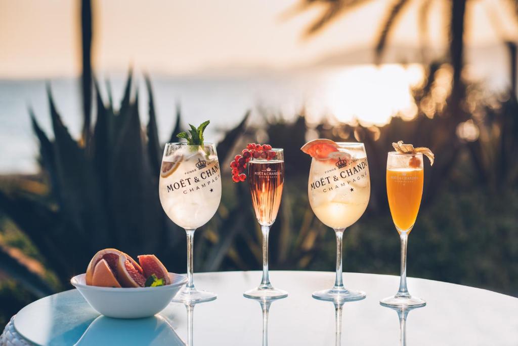 Eleganti dzērieni ar Moët & Chandon zīmolu izkārtoti uz galda, fona ainava ar palmām un saulrietu. Dzērienos redzami augļi un garšaugi, radot svinīgu atmosfēru.