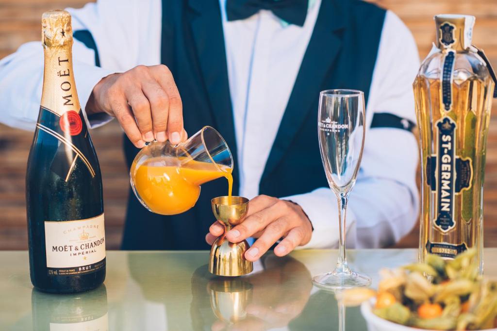 Bārmenis ielej apelsīnu sulu mērīšanas kausā, blakus atrodas šampanietis Moët & Chandon un St-Germain liķieris, radot elegantu kokteiļu sagatavošanas mirkli.