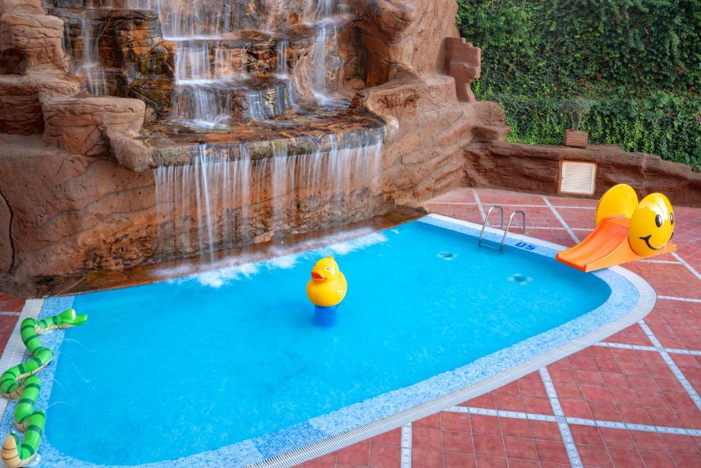 Baseins ar ūdens kritumu un rotaļlietu peldētājiem, piemēram, dzeltenu pīli un slaidu. Apkārtne ir veidota ar dabīgām akmeņu struktūrām un zaļām augu daļām.