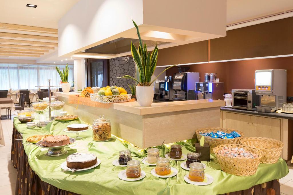 Bagātīgs brokastu bufetes galds ar dažādiem desertiem, augļiem un dzērieniem, kas pasniegti viesnīcā. Zaļais galdauts piešķir viesmīlīgu atmosfēru.