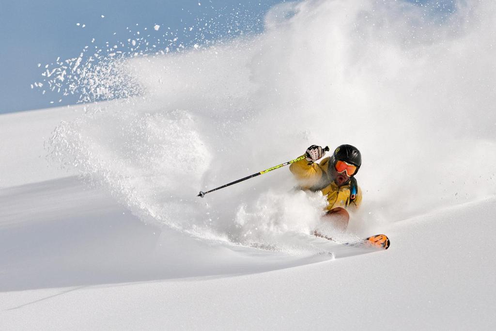 Sniega slēpotājs, kas veic triku pāri dziļam, svaigam sniegam, radot sniega mākoni. Ideāla ainava ziemas sporta entuziastiem.