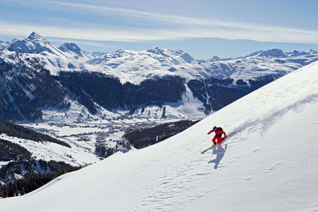 Sniega slēpotājs strauji slīd lejup pa kalnu, apkārt redzami sniegoti kalni un skaista ainava.
