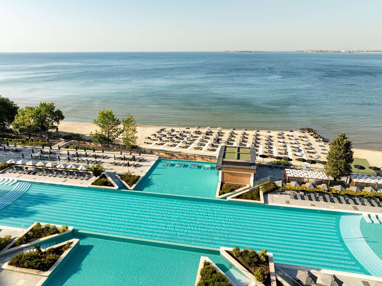 Plašs skats uz pludmali un baseinu, kurā ir daudz sauļošanās krēslu un saulessargu, kas pie jūras. Skats uz mierīgu ūdeni un zilo debesu fonu. panoramic view with beach and pools