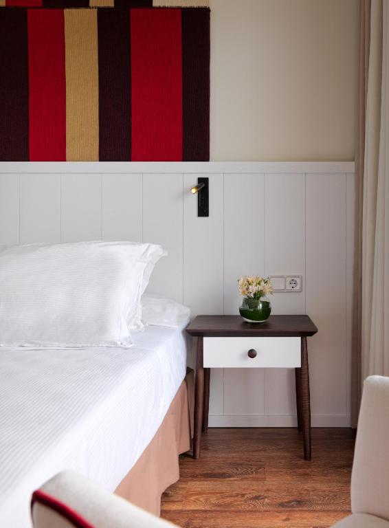 Mūsdienīga guļamistaba ar baltu gultu un koka naktsgaldiņu, uz kura ir puķu pods. Sienā redzama krāsaina auduma kompozīcija.