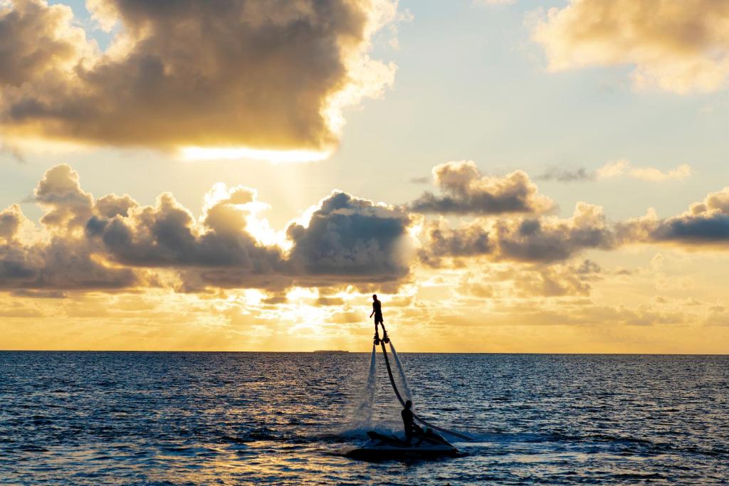 Cilvēks, kurš lidinās virs ūdens, izmantojot ūdenslidojumu, pret saulrietu fona.
