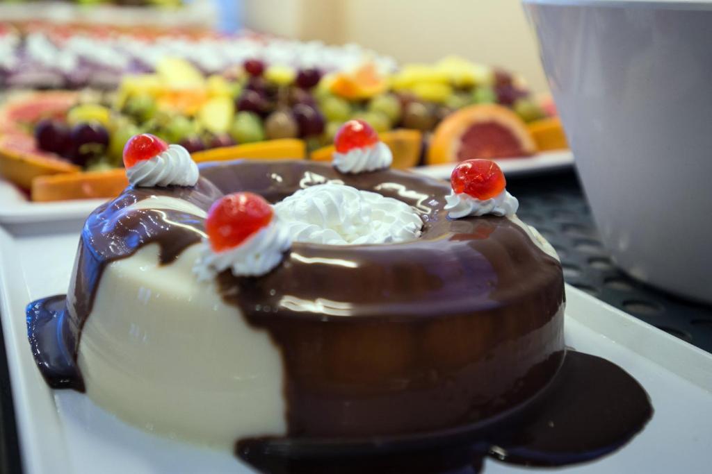 Šokolādes deserts ar saldo krējumu un ķiršiem, pasniegts uz balta šķīvja, fonā redzami augļu izstrādājumi.