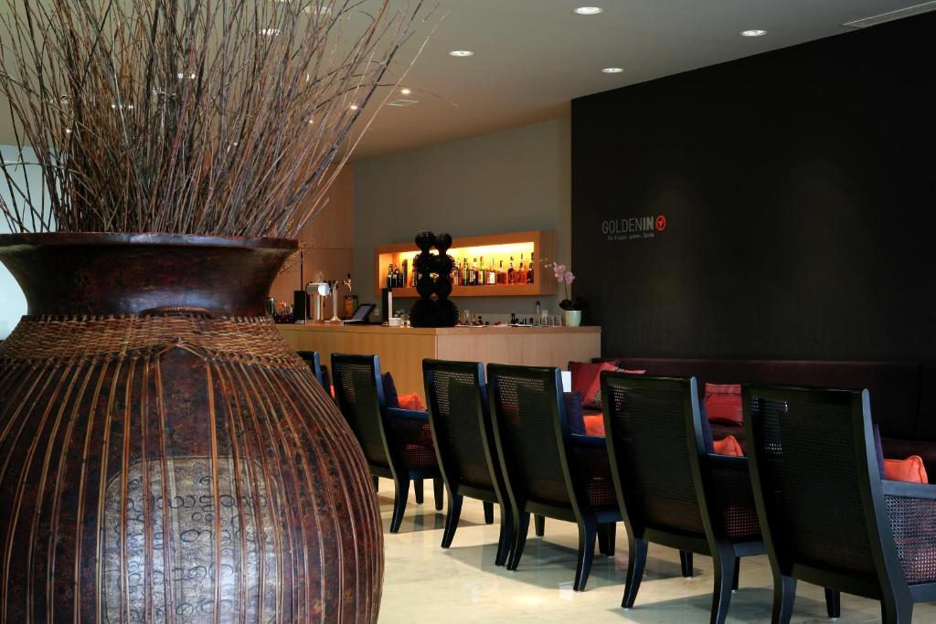 Mūsdienīgs interjers ar lielu dekoratīvo trauku un krēsliem, kas veido relaksējošu atmosfēru. Aiz bāra letes redzama elegantā apgaismojuma zona ar alkoholiskajiem dzērieniem.