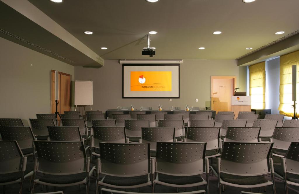 Konferences telpa ar rindu sēdvietām, projektoru un ekrānu, kurā redzams zīmols "GOLDEN SOURCE". Gaisma ienāk caur logiem ar dzeltenām žalūzijām.