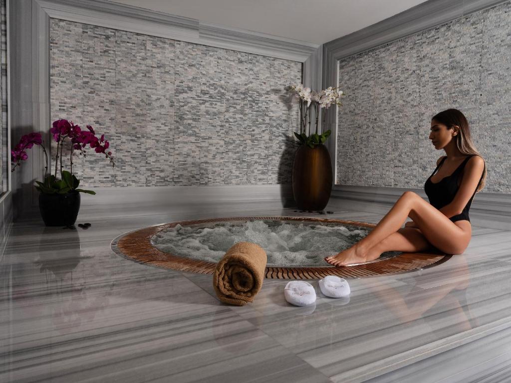 Mūsdienīgs spa interjers ar relaksējošu burbuļvannu, ap kuru sēž sieviete un ir redzami ziedi un dvielis.