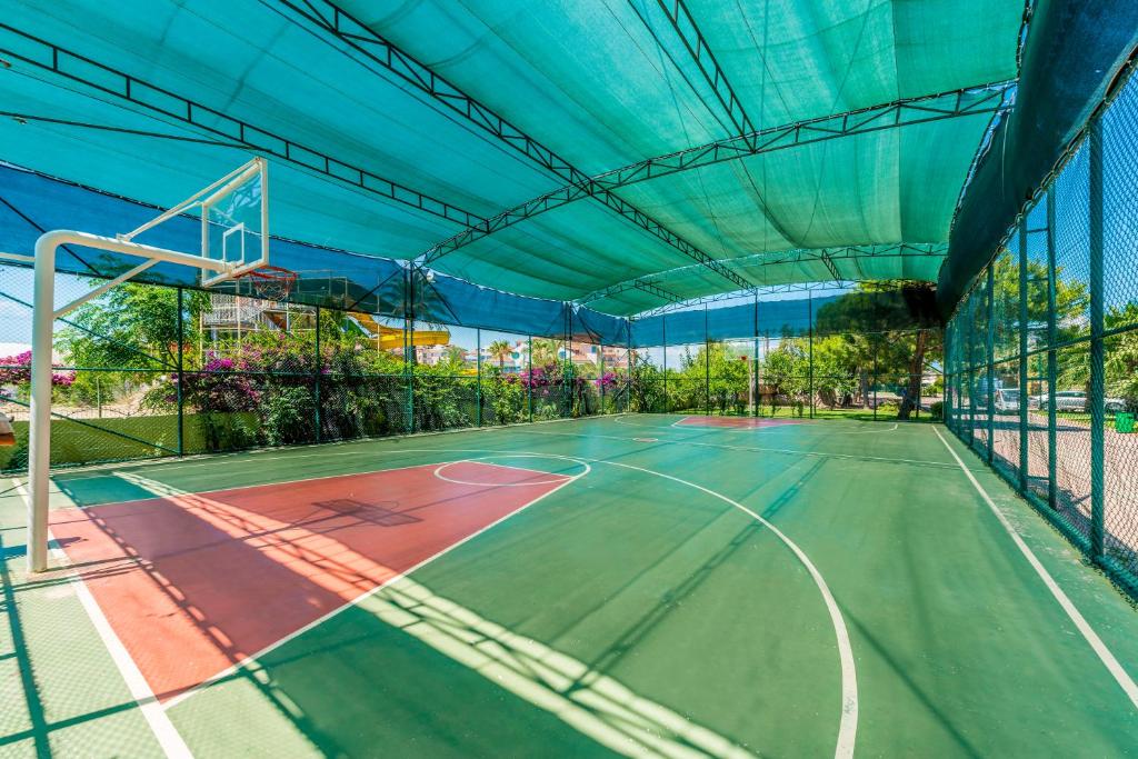 Basketbola laukums ar jumtu, kas nodrošina ēnu, un zaļo un sarkano krāsu. Apkārt ir ziedi un koki, radot patīkamu vidi sportošanai.