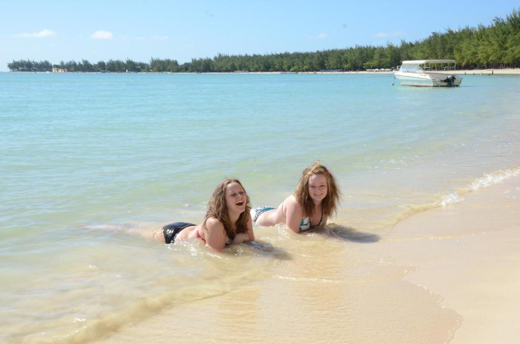 Divas meitenes spēlējas seklajā jūrā, smejoties un baudot saulainu pludmali ar palmām fona. Merces laiva ir redzama tuvumā, bet debesis ir skaidras un zilas.