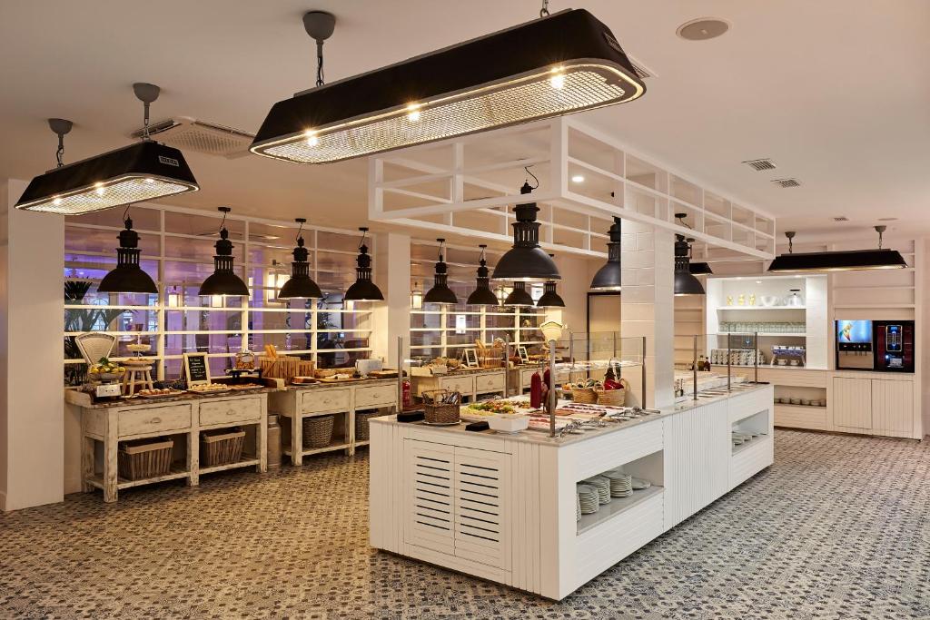 Mūsdienīgs un stilīgs bufetes restorāns ar baltām mēbelēm un melnām lampām, kas rada patīkamu atmosfēru. Galds ir pilns ar dažādiem ēdieniem un dzērieniem, nodrošinot lielisku izvēli viesiem.