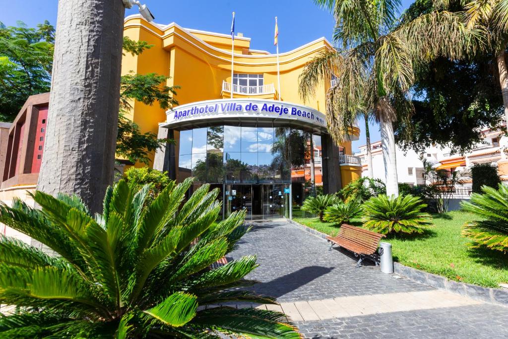 Aparthotel Villa de Adeje Beach ieejas zona ar modernu arhitektūru un zaļo apstādījumu, kas rada viesmīlīgu atmosfēru. Apkārt ir palmām un citiem augiem, kas uzsver tropisko vidi.