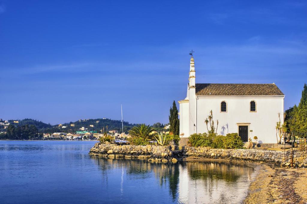Mierinājoša skats uz baltu baznīcu, kas atrodas pie ūdenskrātuves, ar kalniem un pilsētu fona. Zilais debesis un saules gaisma piešķir ainavai mierīgu atmosfēru.