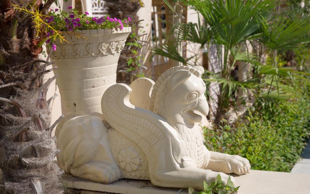 Statuete, kas attēlo leģendāru radījumu ar lauvas galvu un putna ķermeni, atrodas dārzā blakus ziedu podam un palmām. Daba un arhitektūra harmoniski apvienojas, radot mierīgu atmosfēru.