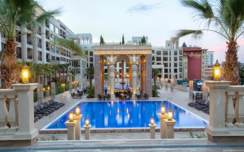 Moderns kūrorta viesnīcas peldbaseins ar palmām un elegantiem apgaismojuma elementi, kas atrodas pie baseina. Apkārt esošās terases ar sauļošanās krēsliem un kafejnīcām rada relaksējošu atmosfēru.