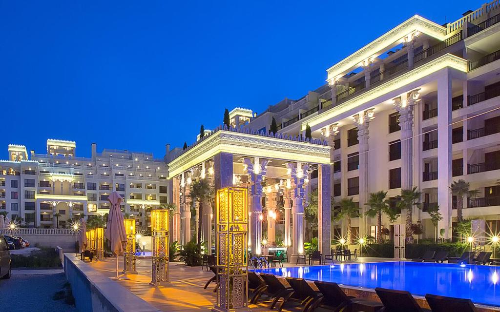 Luksusa viesnīcas komplekss ar elegantu arhitektūru un apgaismotu peldbaseinu, kas izskatās īpaši pievilcīgi tumsā. Zilā debess fonā izceļas moderni ēku elementi un zaļie stādījumi.