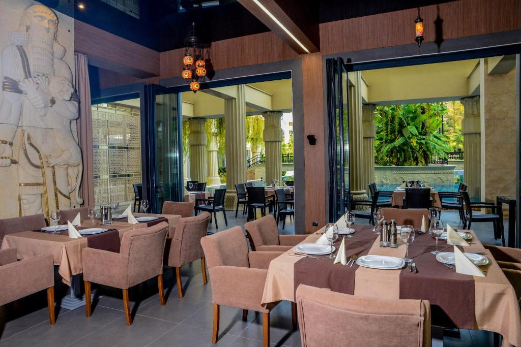 Mūsdienīgs restorāns ar elegantiem galdiem un krēsliem, dekorēts ar greznu sienu skulptūru un ar skatu uz zaļo dārzu.