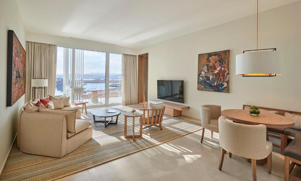 Mūsdienīgs viesistabas interjers ar plašiem logiem, kas sniedz skatu uz ūdeni, un stilīgām mēbelēm, tostarp dīvānu, televizoru un ēdamgaldu.