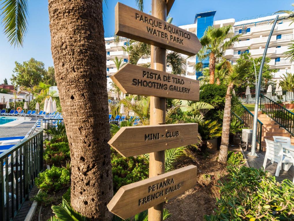 Koka norādes zīme, kas norāda uz dažādām izklaides iespējām, piemēram, ūdens parku, šaušanas galeriju, mini klubu un petanque laukumiem, iestādīta zaļā teritorijā ar baseinu un palmām.