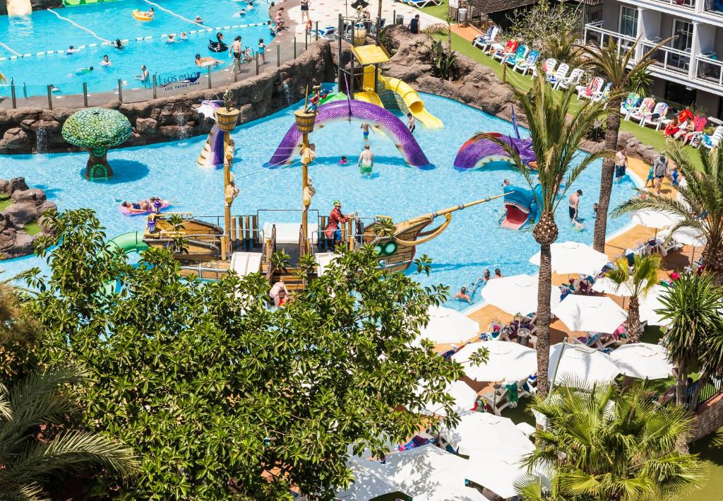 Ūdens atrakciju parks ar kuģa struktūru un slidkalniņu, ap ko pulcējas apmeklētāji. Apkārt baseinam redzami palmām un atpūtas krēsliem.