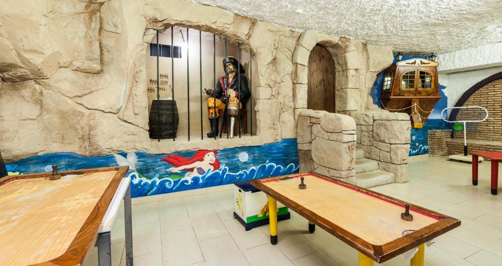 Bērnu spēļu istaba ar jūras pirātu tēmu, kurā ir krāsainas sienas gleznas un spēļu galdi. Telpā ir redzams pirāts aiz restēm un ilustrācija ar mermaid.