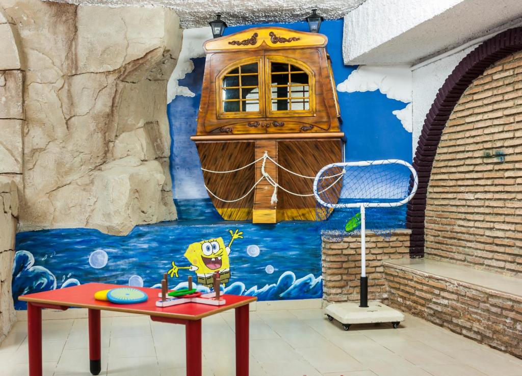 Krāsains interjers ar Spongebob tēlu un kuģa attēlu uz sienas, kas rada jūras atmosfēru. Telpā ir galds ar rotaļlietām un sporta aprīkojumu.