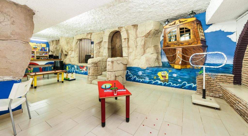 Krāsains bērnu spēļu istaba ar akmens sienām un Spongebob tēlu uz sienas. Telpā ir galdi un krēsli, kas paredzēti radošām aktivitātēm.