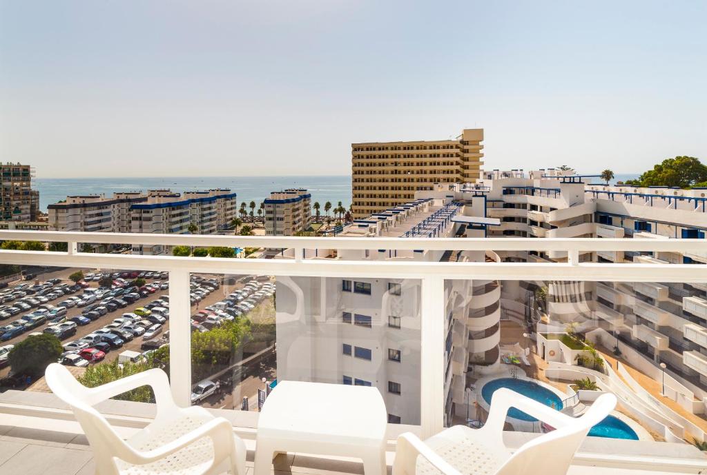 Skats no balkona uz jūru un apkārtējām ēkām, kur redzami daudzdzīvokļu nami un stāvvietas. Gaisma un skaidrs laiks rada patīkamu atmosfēru.