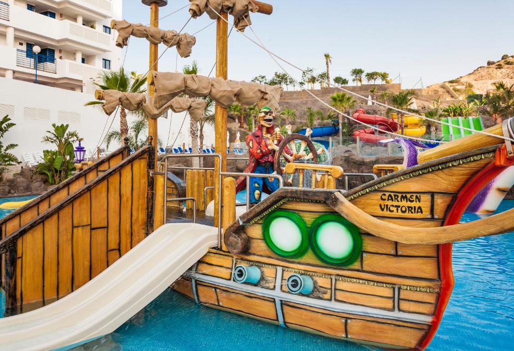 Peldbaseins ar bērnu slidkalniņu un krāsainu kuģi, kas atrodas ūdenī, nodrošinot jautru un aizraujošu vidi ģimenēm. Apkārt redzamas palmas un citi ūdens atrakciju elementi.