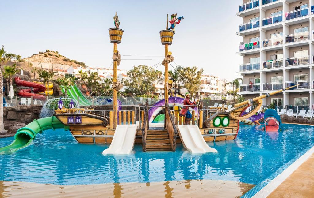 Bērnu ūdens atrakciju parks ar pirātu tēmu, kas ietver slidkalniņus un rotaļu kuģi, apkārt palmām un viesnīcas ēku fona. Ideāli piemērots ģimenēm un bērniem.