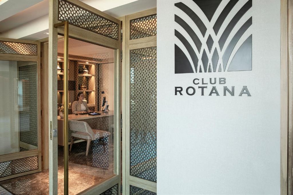 Skaista un moderna reģistratūra "Club Rotana" ar elegantu dekoru un siltu apgaismojumu, kur darbiniece strādā pie datora.