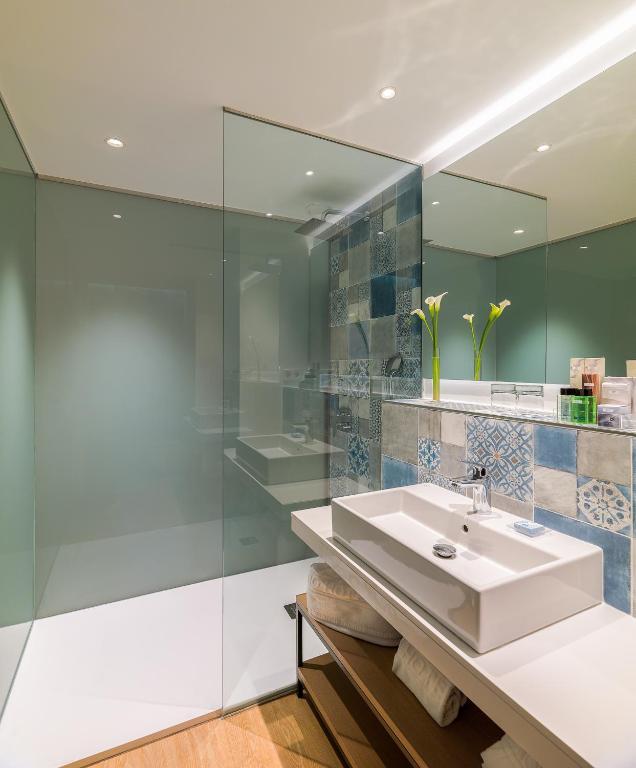 Mūsdienīgs vannas istabas interjers ar stikla sienām, stilīgu izlietni un krāsainām flīzēm. Telpa ir gaiša un eleganta, ar minimālistisku dizainu un dekoratīvām ziedu kompozīcijām.