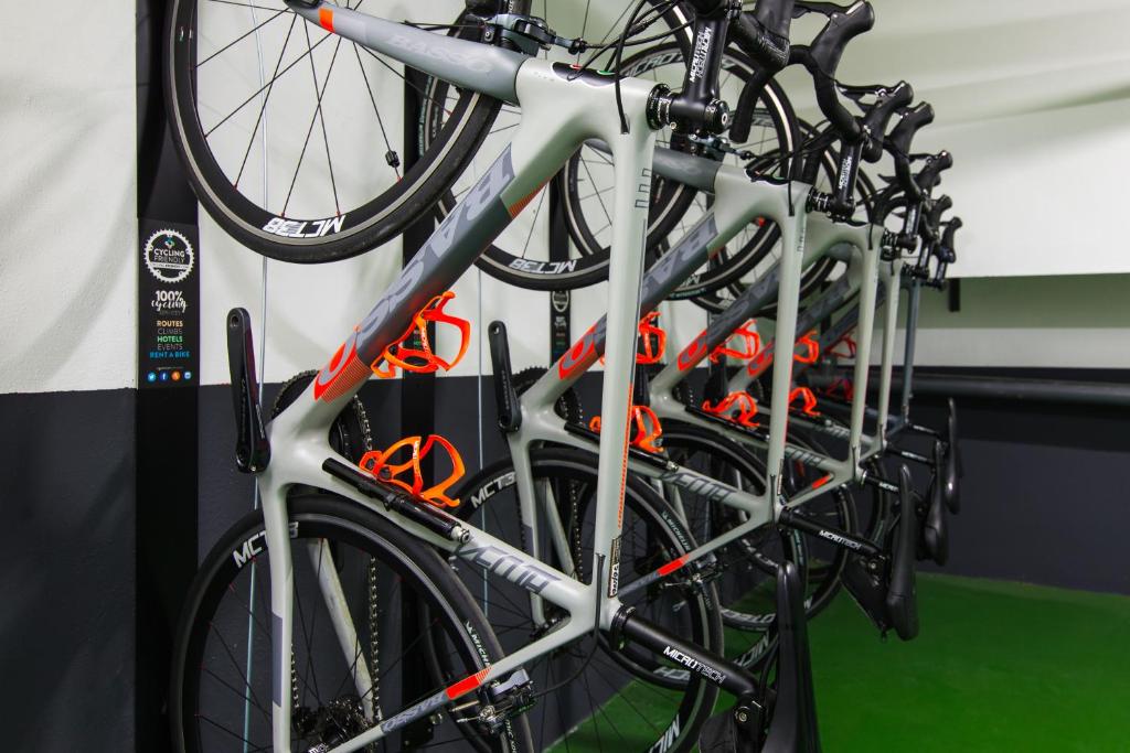 Velosipēdu stends ar vairākiem moderniem velosipēdiem, kas sakārtoti uz sienas. Velosipēdiem ir pelēka un oranža krāsa, kas izceļ to sportisko dizainu.