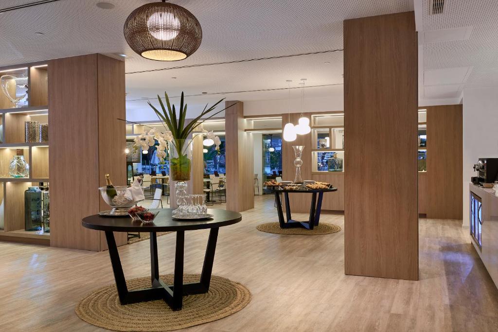 Eleganta viesnīcas uzgaidāmā telpa ar modernu dizainu, kurā ir stilīgi galdi, dekoratīvi elementi un apgaismojums. Telpā ir redzami ziedi un dzērieni, radot viesmīlīgu atmosfēru.