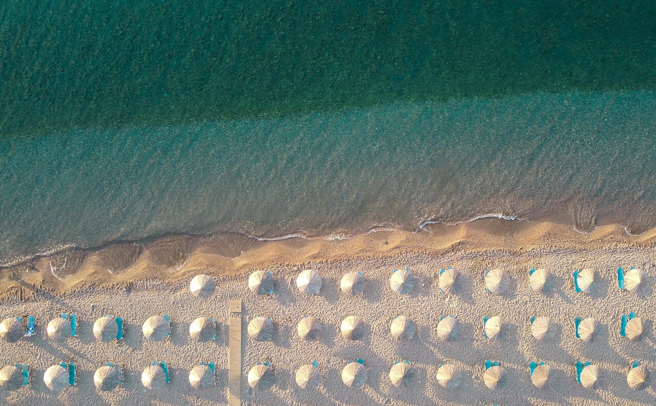 Skats no augšas uz Mitsis Rodos Maris privāto smilšu pludmali ar tirkīzzilu ūdeni un saulessargiem.