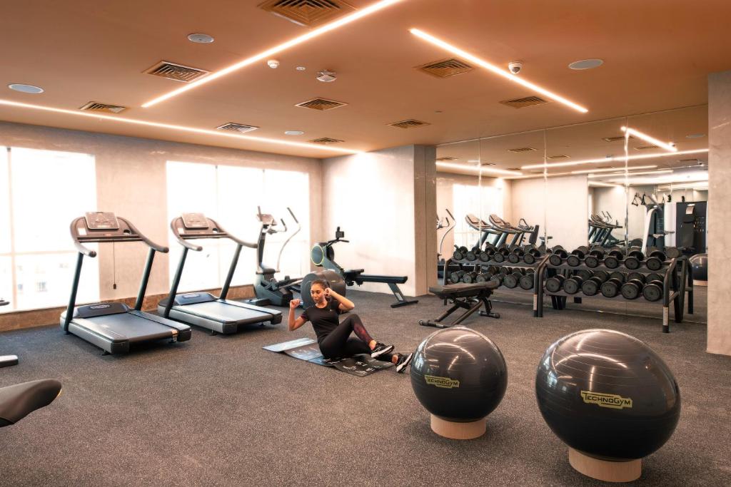 Mūsdienīga trenažieru zāle ar dažādām sporta iekārtām, tostarp skrejceliņiem, velosipēdiem un hantelēm, kā arī vingrošanas bumbām. Sieviete veic vingrojumus uz paklāja, radot aktīvas dzīvesveida atmosfēru.