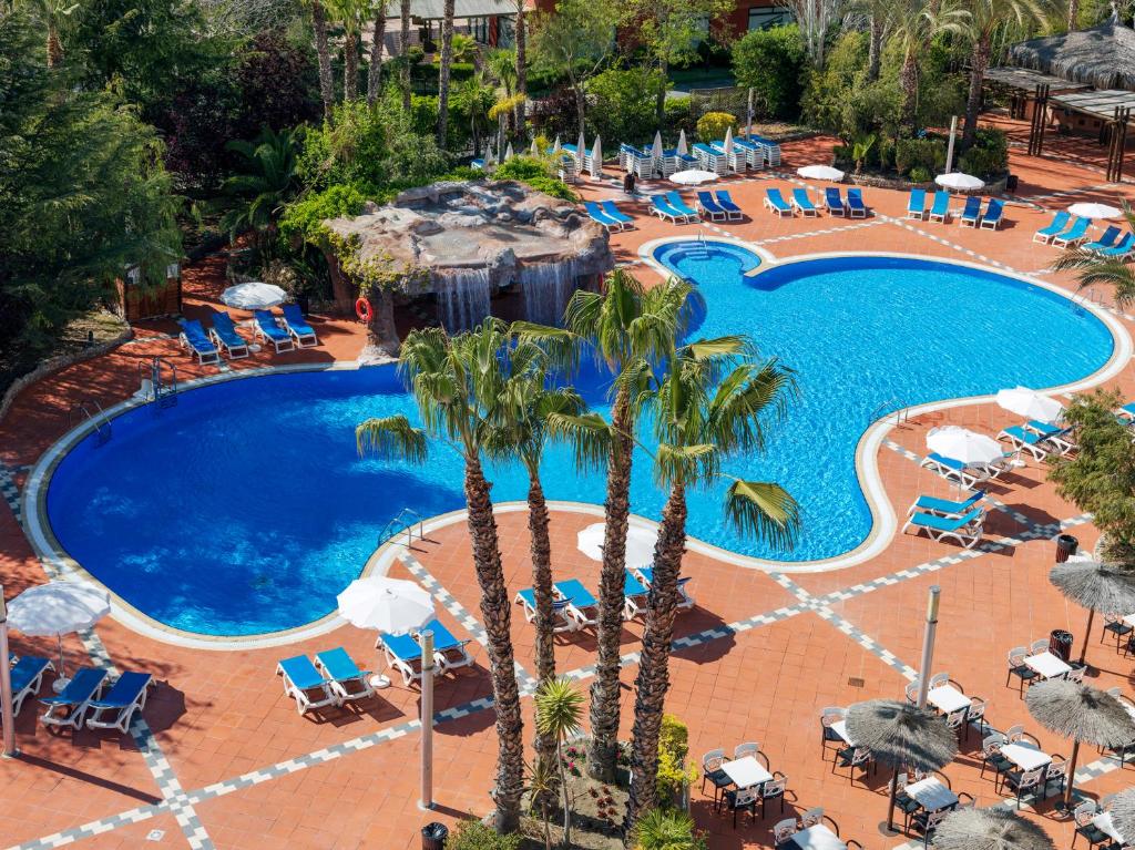 Lielisks skats uz viesnīcas baseinu ar zilu ūdeni, palmu kokiem un sauļošanās krēsliem. Baseins ir veidots kā peldēšanas zona un ir apzaļumots ar krāšņiem augiem.