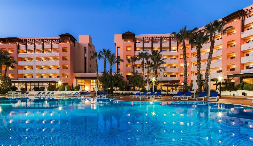 Luksusa viesnīca ar modernu arhitektūru un peldbaseinu, apstādīta ar palmām, kas izgaismota vakara gaismā.