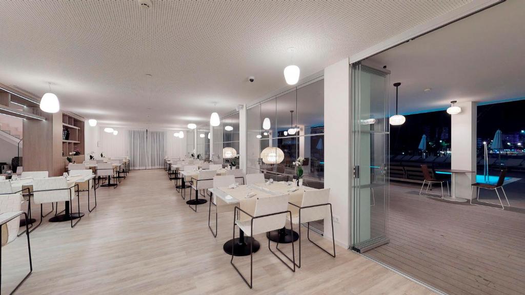Mūsdienīgs restorāna interjers ar gaišām, eleganti iestatītām galdiem un stikla durvīm, kas ved uz terasi pie baseina. Apgaismojums radīšanai patīkamu atmosfēru.