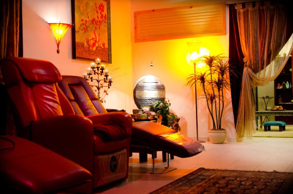 Relaksējoša telpa ar sarkaniem masāžas krēsliem, siltu apgaismojumu un augiem, radot mierīgu atmosfēru. Sienas rotā glezna, bet stūrī ir dekoratīvs ūdens elements.