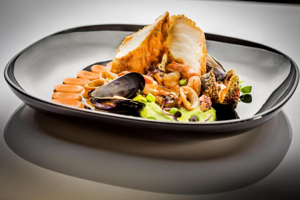 Elegants kalnu ēdiens, kas sastāv no garnelēm, mīdijām un zivs filejas, pasniegts uz melnas šķīvja ar krāsu kontrastiem un garnējumu.