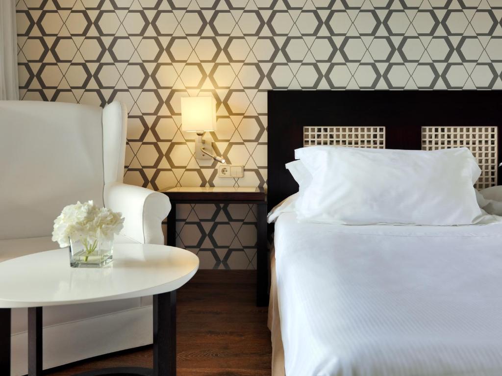 Mūsdienīgs guļamistabas interjers ar elegantu tapetu, ērtu krēslu un gultu ar baltām spilveniem, kas papildināts ar ziedu kompozīciju uz galda.