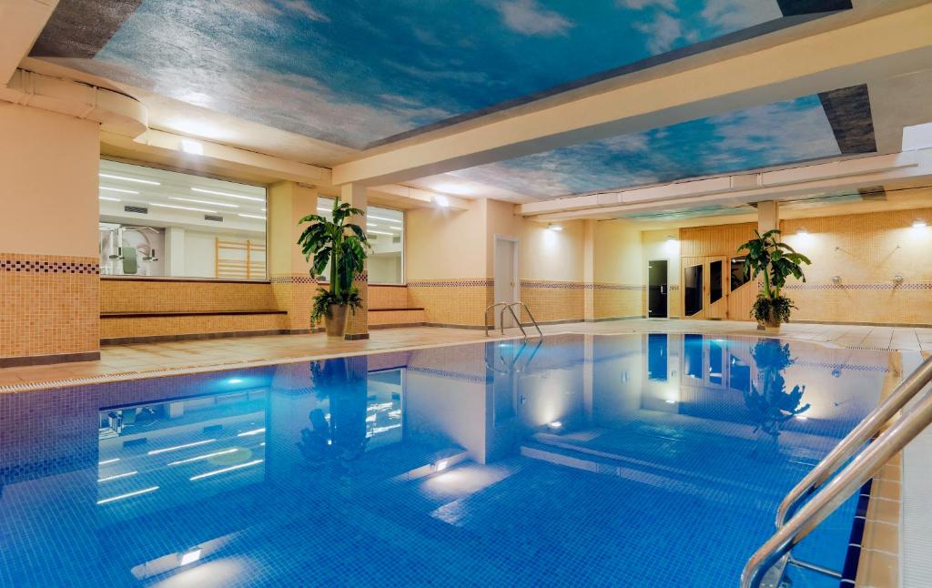 Iekštelpu peldbaseins ar zilu ūdeni un debesu motīvu griestiem, apkārt peldbaseinam ir augi un sporta aprīkojums.