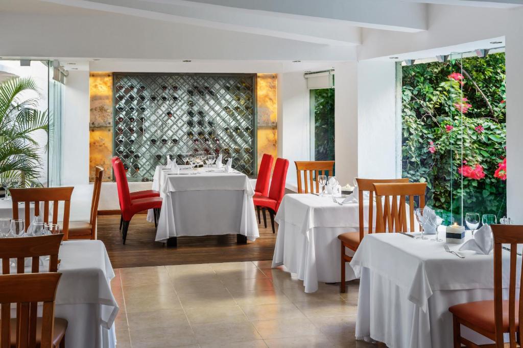 Eleganta restorāna interjers ar baltām galdautām un sarkaniem krēsliem, redzami ziedi caur stikla sienām. Gaisīga atmosfēra ar vīna plauktu un dabu fonā.
