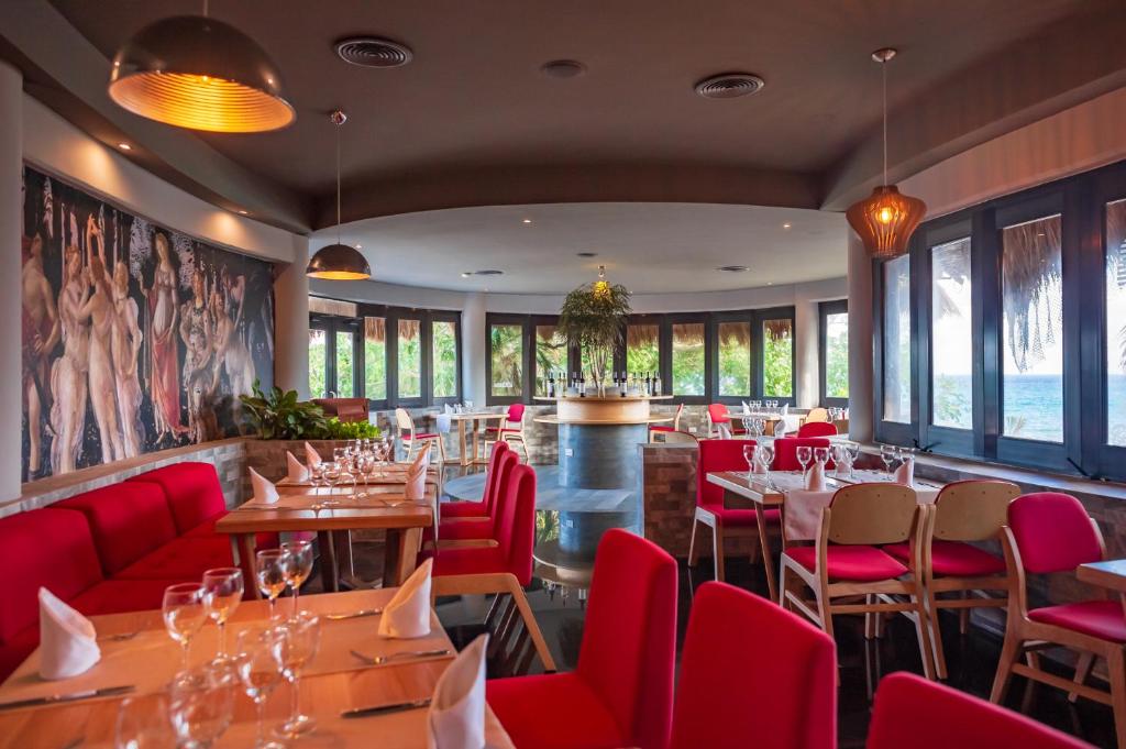 Mūsdienīgs restorāna interjers ar sarkaniem krēsliem, koka galdiem un gleznainu sienu, kas rotāta ar mākslas darbu. Lieliski skati uz jūru, kas redzami caur lielajiem logiem.
