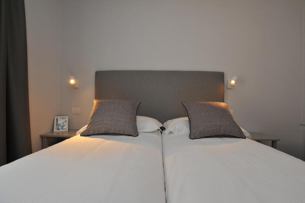 Mūsdienīga guļamistabas interjers ar divām atsevišķām gultām, uz kurām ir dekoratīvās spilveni. Sienas apgaismojums un stilīgs naktsgaldiņš ar foto rāmi papildina telpas dizainu.