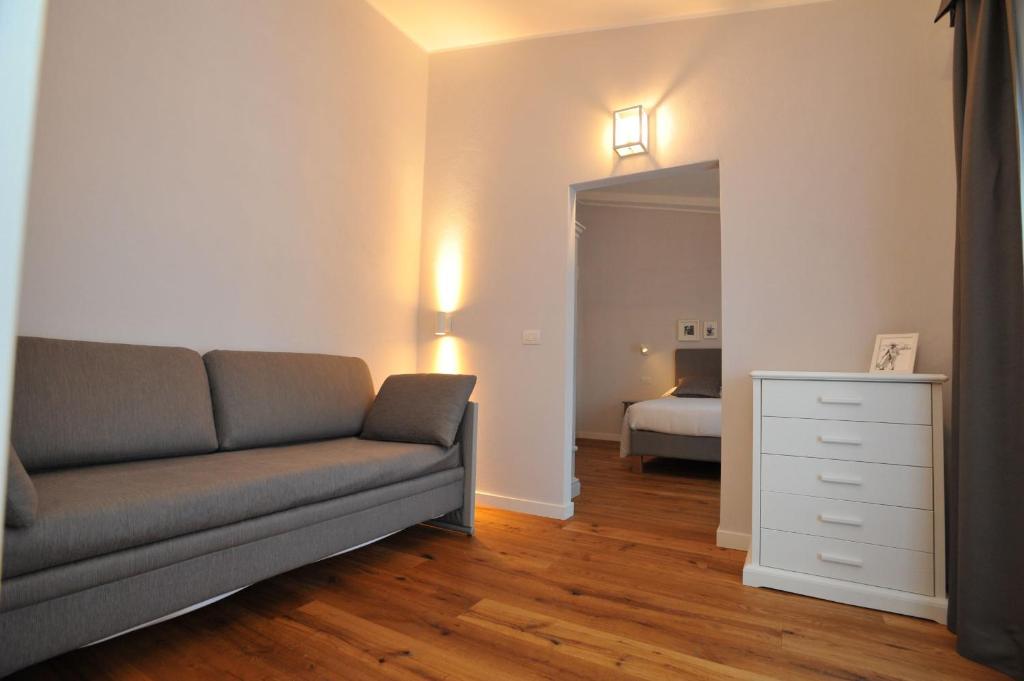 Gaiša un moderna viesistaba ar pelēku dīvānu un baltu kumodi, redzama durvju atvere uz guļamistabu. Koka grīda piešķir siltumu telpai.