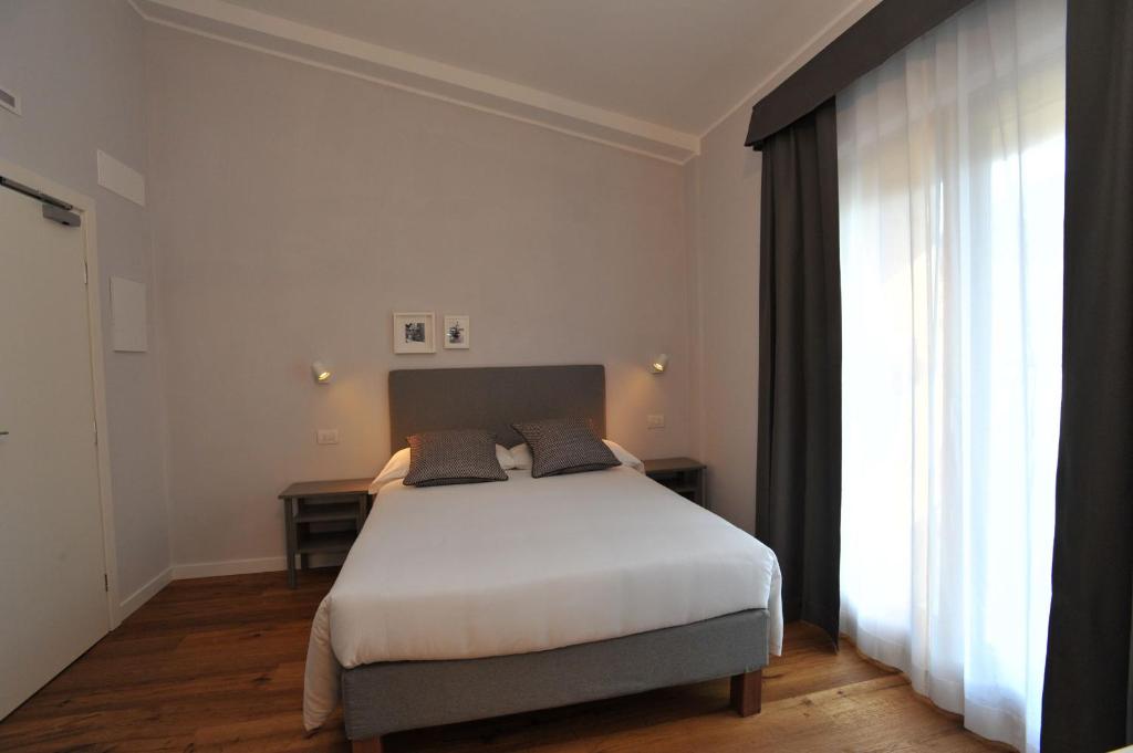 Mūsdienīga guļamistaba ar ērtu gultu, koka grīdu un lieliem logiem, kas nodrošina dabisko apgaismojumu. Sienas ir gaiši pelēkas, un telpā ir minimālistisks dekors ar dažām fotogrāfijām.