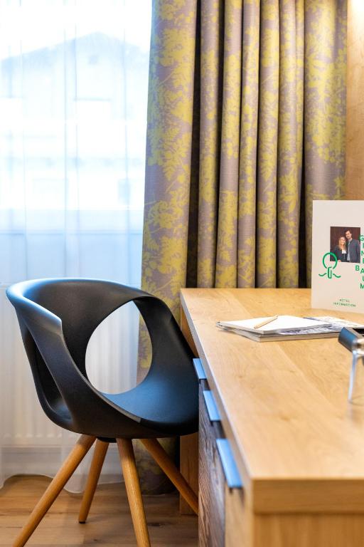 Moderns rakstāmgalda stūris ar melnu krēslu, koka virsmu un rotājošām aizkariem. Uz galda atrodas piezīmju bloks un apdrukāta lapa.